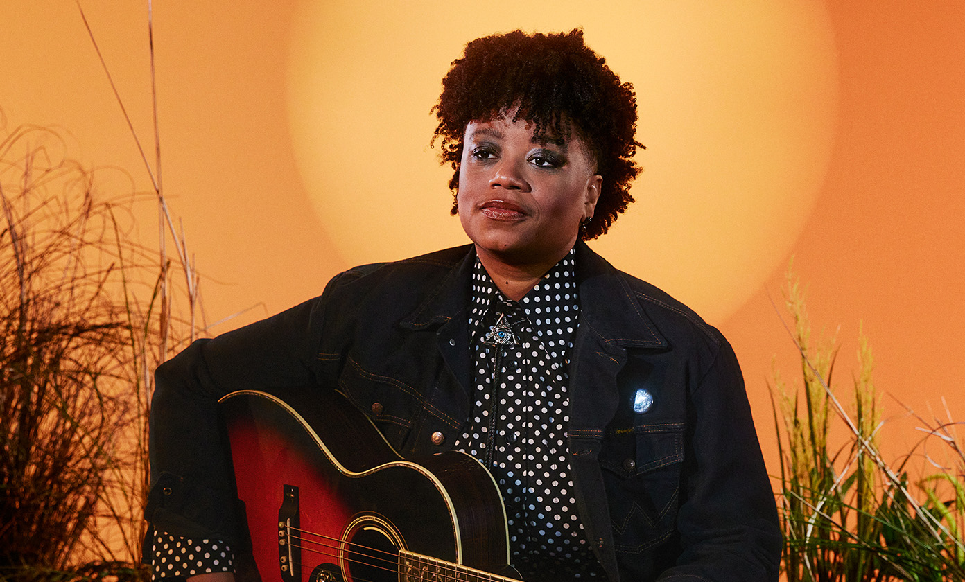 Amythyst Kiah looking off to the side sitting on a stool with her guitar